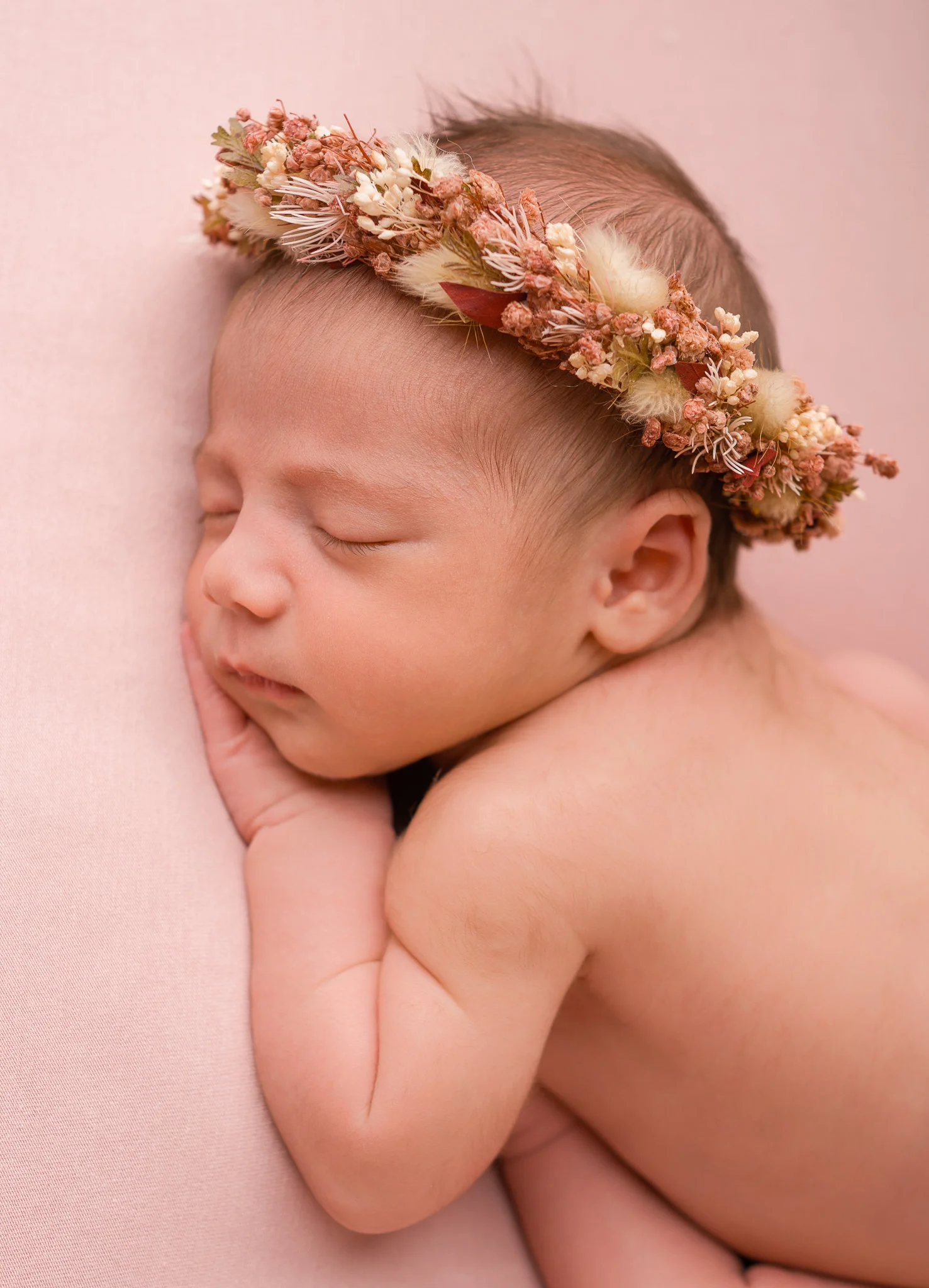 Sovende nyfødt baby med blomsterkrans på hodet, liggende på en myk rosa bakgrunn under profesjonell nyfødtfotografering i studio.