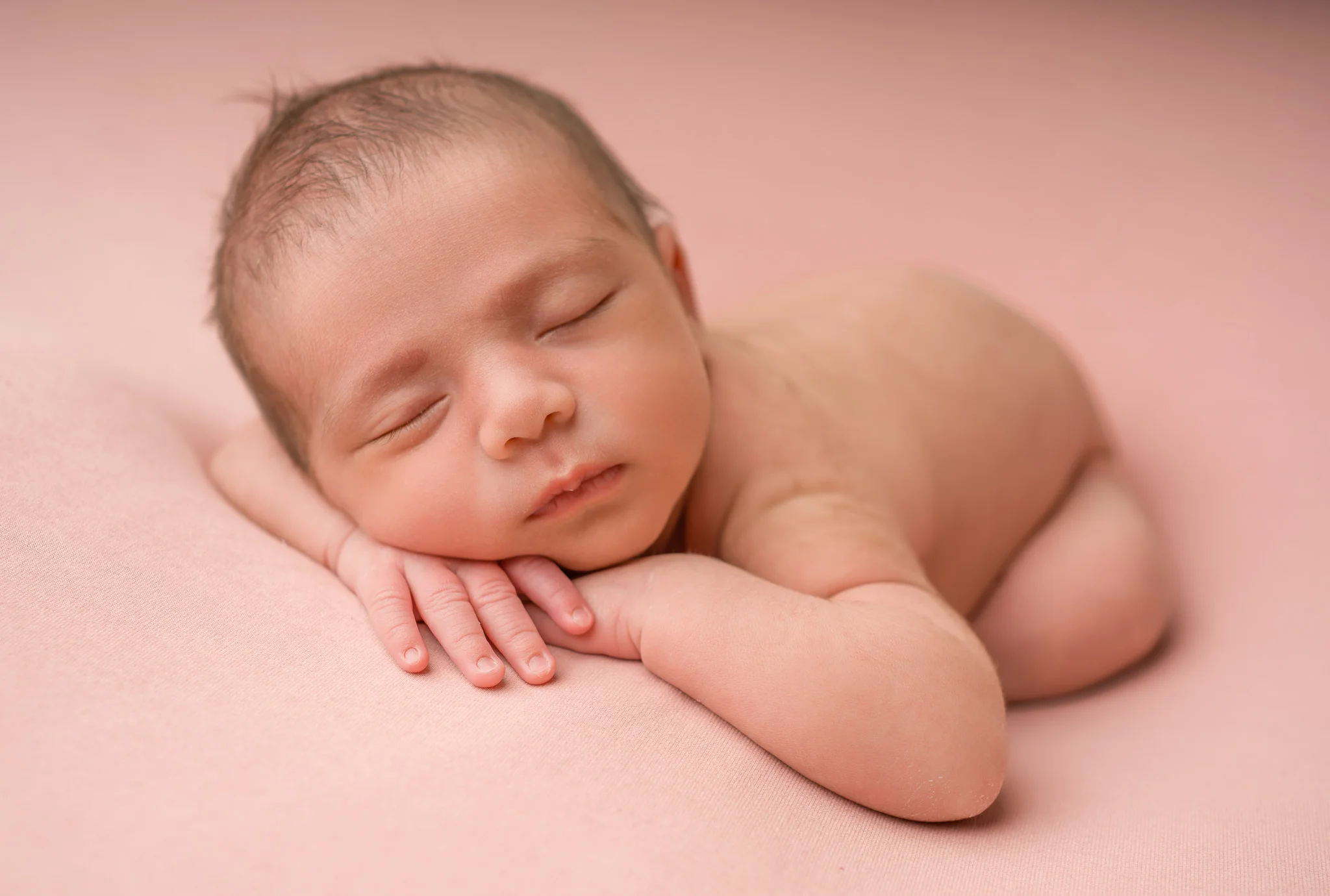 Nyfødt baby sover fredelig på magen med hendene under kinnet på et mykt, støvrosa underlag i et profesjonelt fotostudio.