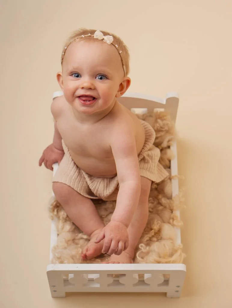 Smilende baby med beige bleiebukse sitter i en liten hvit seng fylt med myk ull under profesjonell babyfotografering.