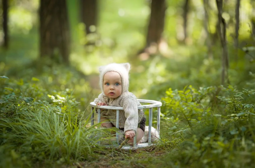 En liten baby med strikket bjørnelue sitter i en rund, hvit trekasse i skogen under utendørs babyfotografering.